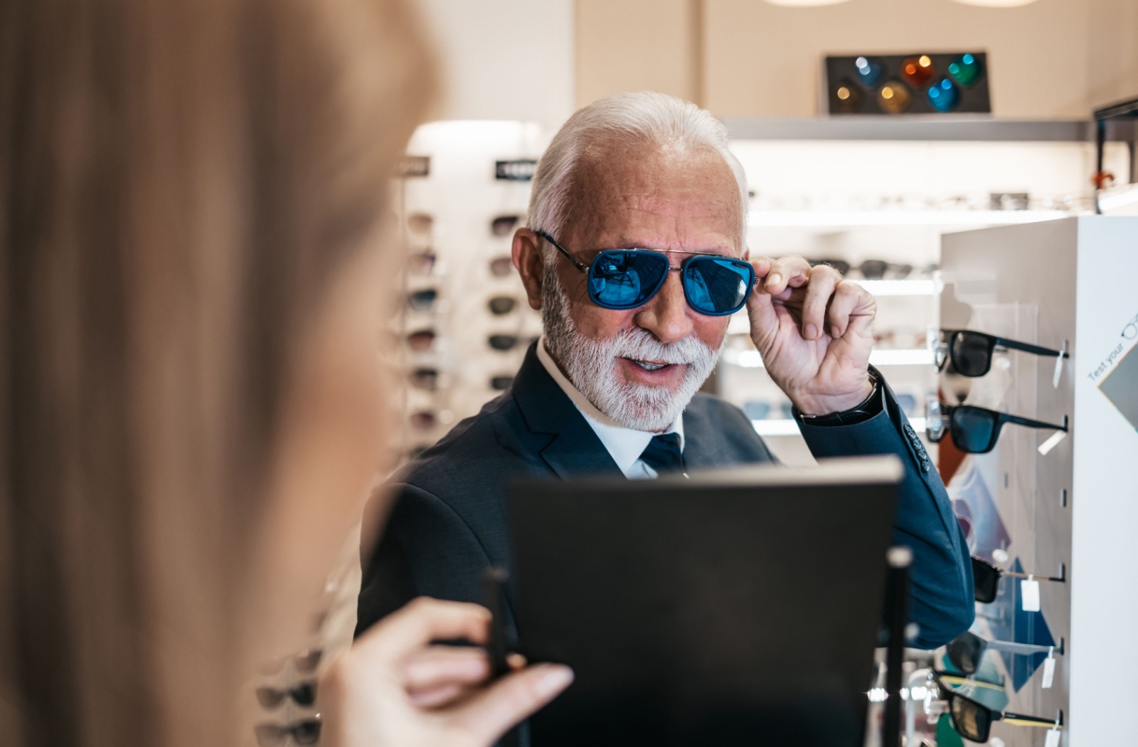 Elegant senior man tries on sunglasses while eye care staff member holds up a mirror.