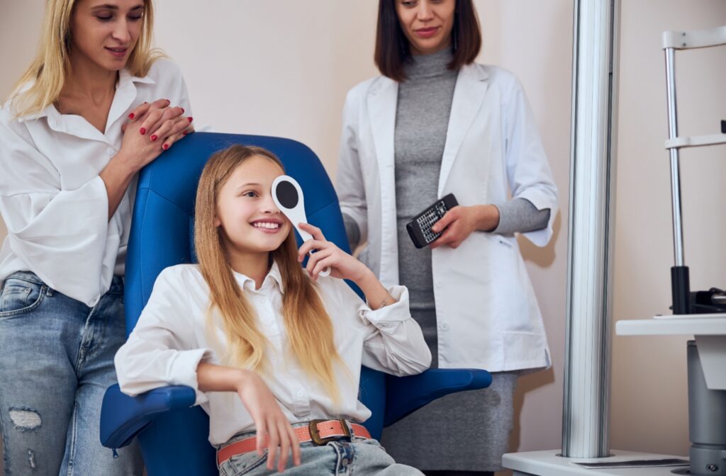 A child covers 1 eye during an eye exam with their parent and optometrist standing behind them.