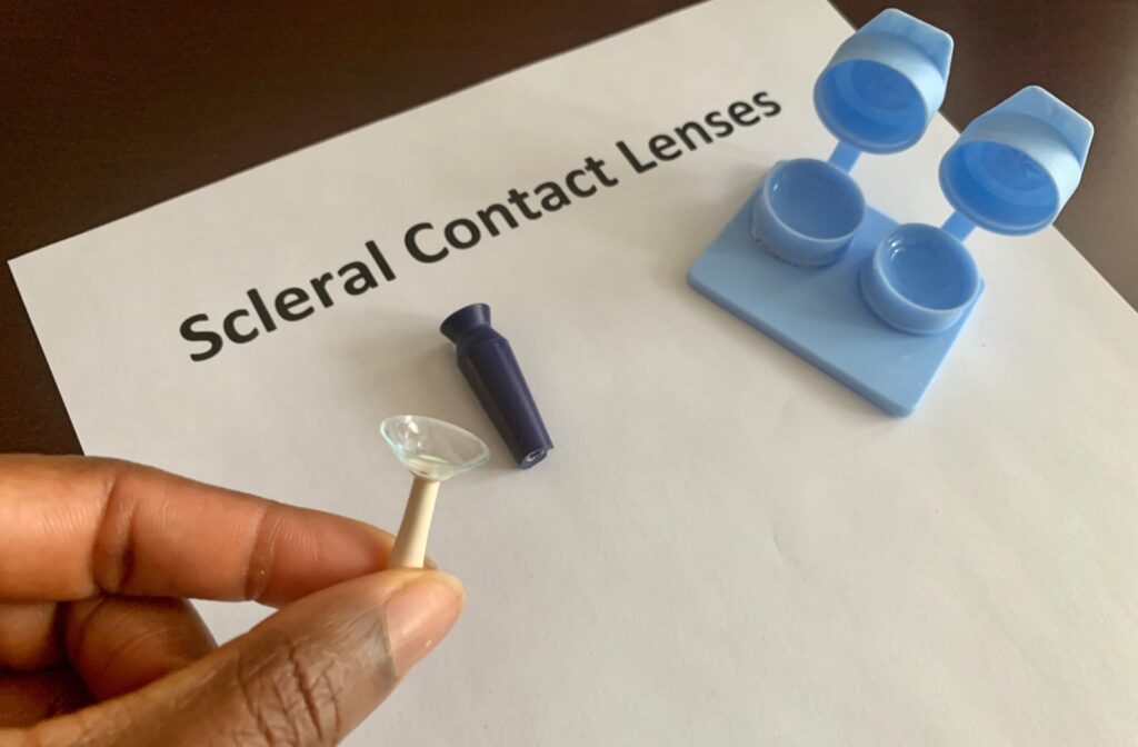 Fingers holding an applicator with a scleral lens, with "Scleral Contact Lenses" on a piece of paper in the background along with other accessories.