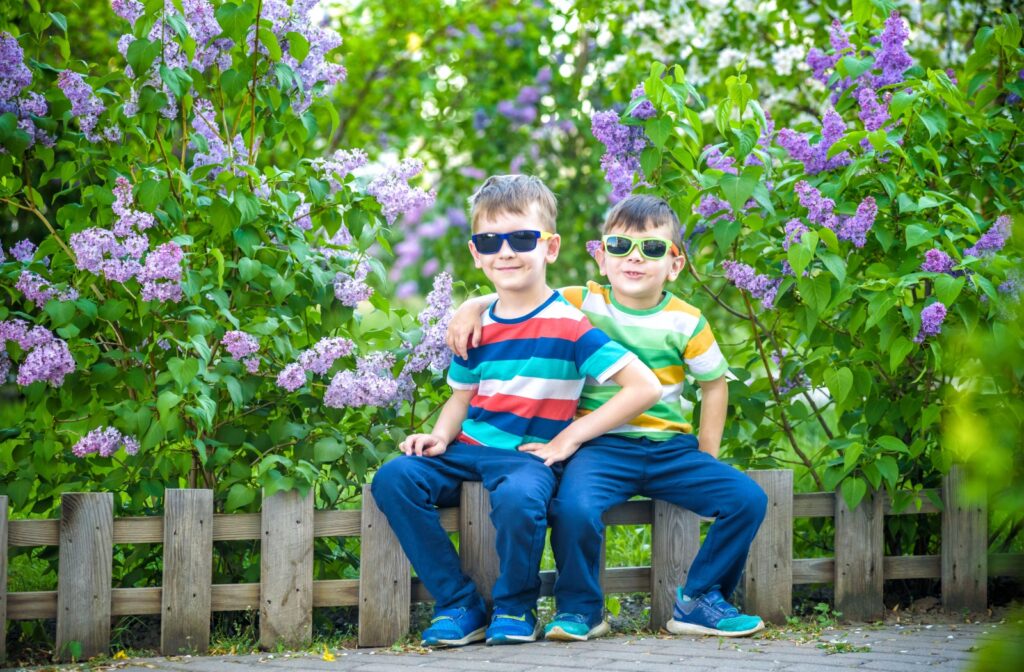 2 young children sitting on a low fence outdoors and wearing sunglasses.