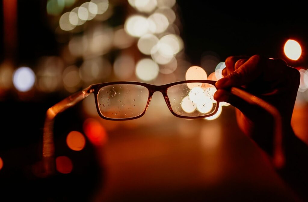 Person holding eyeglasses up at night with blurred city lights in the background, showing raindrops on the lenses and a bokeh effect that illustrates unclear vision or light distortion.