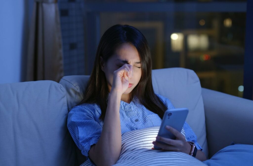 A person sitting on a couch at night rubbing her eyes while looking at her smartphone, illuminated by the screen’s blue light, illustrating eye strain or discomfort from prolonged screen use.