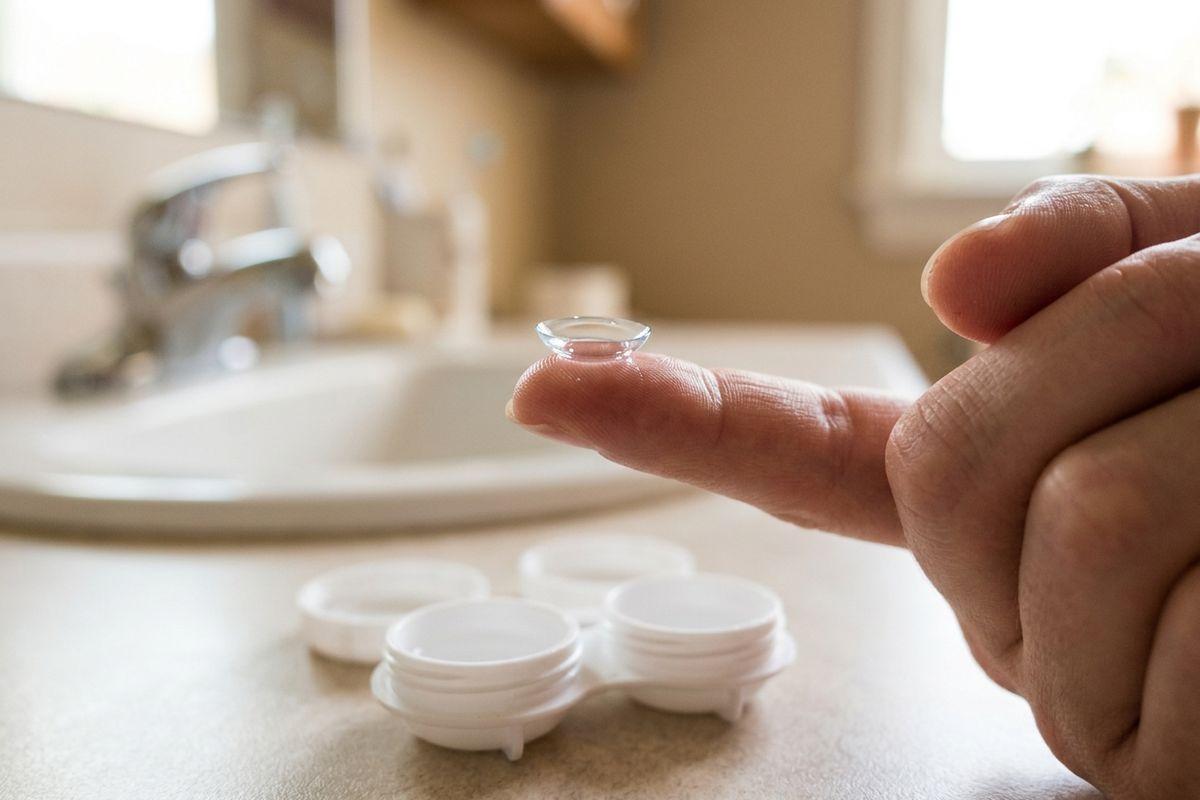 Fingertip holding a clear contact lens above an open white lens case on a bathroom counter, with a sink and faucet blurred behind.